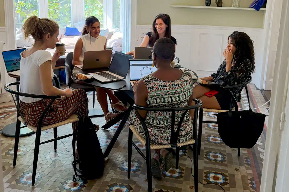 Five women sitting around a table with open laptops and coffee in a bright room with tall windows and a patterned tiled floor